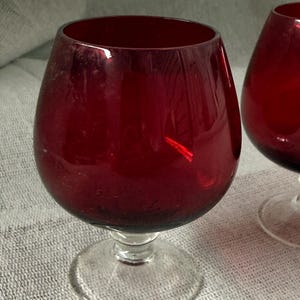 Può includere: Due calici di vino rosso rubino con una coppa arrotondata e uno stelo trasparente. I calici sono in vetro e poggiano su una superficie strutturata. I calici sono vuoti.