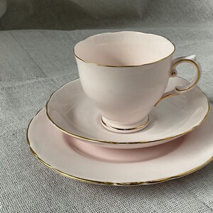 Könnte beinhalten: Ein dreiteiliges Teeservice in zartem Rosa mit goldenen Verzierungen. Das Set besteht aus einer Teetasse, einer Untertasse und einem Kuchenteller. Die Tasse hat einen filigranen Henkel und einen gewellten Rand. Auch die Untertasse und der Teller haben gewellte Ränder.