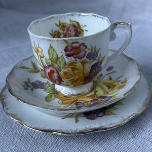 May include: A vintage tea set comprising a teacup, saucer, and side plate. The white porcelain has a floral pattern of yellow, purple, and red roses. The edges are scalloped and trimmed with gold.