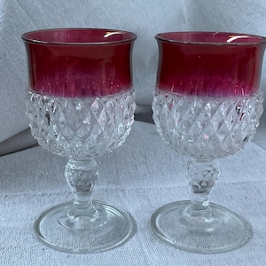 Puede incluir: Dos copas de vino antiguas con cuencos de color rojo rubí y bases transparentes con un patrón de corte de diamante. Los tallos y las bases circulares son de vidrio transparente. Las copas están sobre una superficie clara y texturizada.