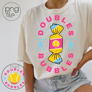 May include: A white t-shirt featuring a pink and blue graphic of a sweet with a softball inside. The text "Doubles & Bubbles" is written around the sweet graphic.