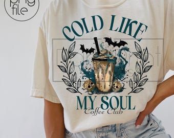 May include: An off-white t-shirt featuring a graphic of a coffee cup with blue liquid, bats, and skulls. The text reads COLD LIKE MY SOUL Coffee Club in dark teal lettering. The design is framed by two vertical leafy branches on each side.