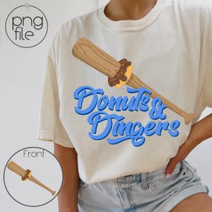 May include: Cream-colored t-shirt featuring a baseball bat with a donut graphic. The donut is brown with sprinkles, and the bat is light brown. The text "Donuts & Dingers" is in blue script.