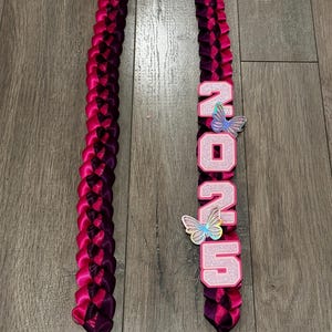 May include: Two braided leis in shades of pink and purple. One lei has the word "2025" in pink glitter letters with butterfly accents. The leis are made of satin ribbon and are displayed on a wooden surface.