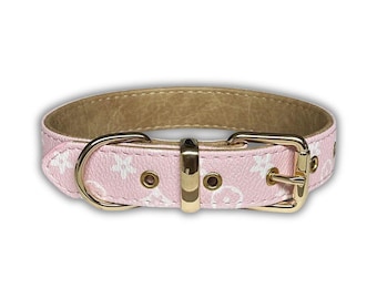 May include: Pink dog collar with a white floral pattern and gold-colored hardware. The collar features a buckle closure and a D-ring for leash attachment. The interior of the collar is a light brown color.