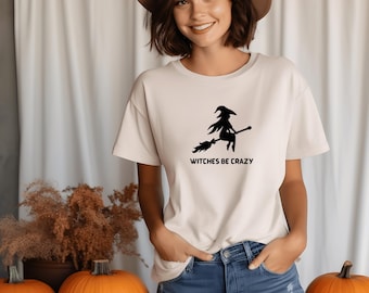 Può includere: Una maglietta color crema con la scritta "Witches Be Crazy" e una silhouette nera di una strega su una scopa. La maglietta è abbinata a jeans blu. Tre zucche arancioni intagliate sono in primo piano, suggerendo un tema di Halloween.