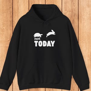 May include: Black hooded sweatshirt with a kangaroo pocket. The front features a white graphic of a turtle and a rabbit, with the text "not TODAY" in white block letters. The background is a wooden surface.