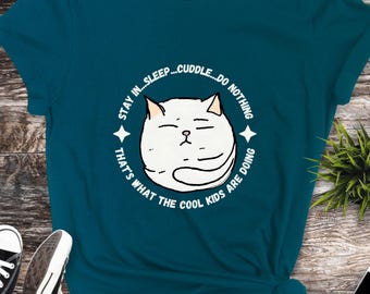 Puede incluir: Camiseta azul turquesa con un gráfico blanco de un gato durmiendo. El gráfico tiene el texto "STAY IN... SLEEP... CUDDLE... DO NOTHING THAT'S WHAT THE COOL KIDS ARE DOING".