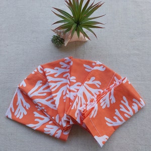 May include: A stack of orange fabric napkins with a white coral reef pattern. The napkins are fanned out, showcasing the design. A small succulent and a decorative plant in a seashell are in the background.