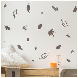 May include: A white wall with brown leaf-shaped wall decals. The decals are arranged in a random pattern and include acorns. A wooden shelf with a candle and a glass jar is in the foreground.