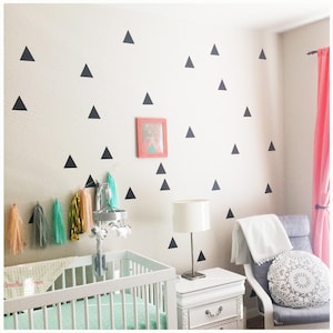 May include: A nursery room with a white crib, a gray rocking chair, and a white nightstand. The wall is decorated with black triangle decals and a framed picture. A mobile hangs above the crib, and colorful tassels are strung along the wall.