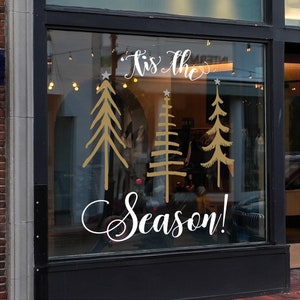 May include: A window decal featuring the white cursive text 'Tis The Season!' with three stylized gold Christmas trees positioned between the words. Each tree is topped with a small white star, creating a festive display on the glass surface.