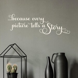 May include: A gray wall with white cursive text that reads "...because every picture tells a Story...". Below the text are black decorative items, including a geometric metal frame with a potted cactus and two black vases.