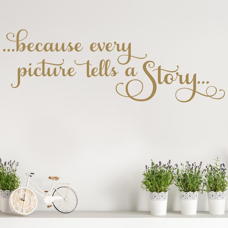 May include: A wall decal in gold script reads "...because every picture tells a Story...". Below, a white bicycle with a clock face for a wheel and potted lavender plants in white pots decorate a shelf against a white wall.