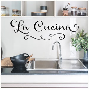 May include: A black and white kitchen wall decal with the text "La Cucina" in a cursive font. The decal is above a stainless steel kitchen sink with a chrome faucet.