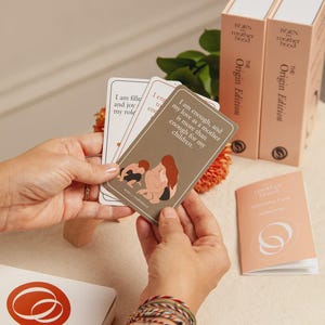 Può includere: Un mazzo di carte di affermazione con il testo "Sono abbastanza, e il mio amore di madre è più che sufficiente per i miei figli". Le carte sono tenute in mano, con due scatole e un libretto sullo sfondo. Le scatole recitano "Born Motherhood, The Origin Edition."