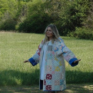 May include: A long, patchwork quilt coat with a variety of colorful fabric squares. The coat has blue cuffs and is worn over a white dress. The background is a green field with trees.