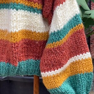 Pu&ograve; includere: Un maglione a righe di maglia con maniche lunghe e una trama a maglia grossa. Il maglione presenta strisce di colore ruggine, bianco, giallo senape e verde acqua.