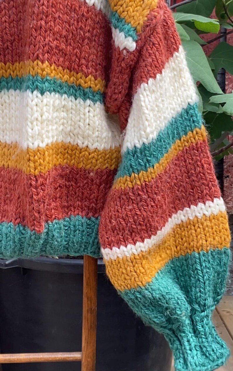 Pu&ograve; includere: Un maglione a righe di maglia con maniche lunghe e una trama a maglia grossa. Il maglione presenta strisce di colore ruggine, bianco, giallo senape e verde acqua.