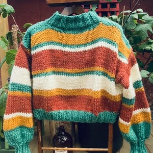 Pu&ograve; includere: Un maglione a righe di maglia con collo alto e maniche lunghe. Il maglione &egrave; realizzato con un filato spesso e presenta strisce alternate di verde, bianco, arancio e marrone.