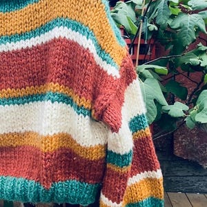 Pu&ograve; includere: Un maglione di lana grossa con un motivo a righe in tonalit&agrave; turchese, bianco, giallo senape e ruggine.