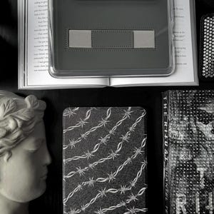 Puede incluir: Una funda de tableta gris oscuro con una banda elástica gris y el texto "Dark And Unhinged". Un libro en blanco y negro con un diseño de alambre de púas está al lado de un busto de mujer.
