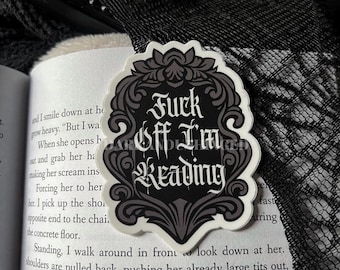 May include: A white sticker with a black and gray ornate border. The sticker reads "Fuck Off I'm Reading" in a gothic font. The sticker is placed on an open book.