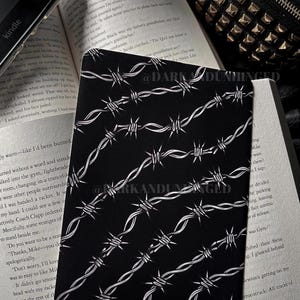 May include: A black rectangular card with a white barbed wire pattern. The card is resting on an open book. The text "@DARKANDUNHINGED" is repeated on the card.