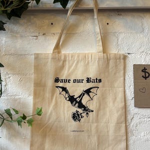 May include: Cream-coloured tote bag featuring a black bat illustration and the text "Save our Bats". The bag has long handles and is suspended against a white brick wall. The bag also has a rose.