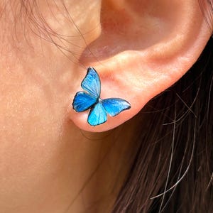 May include: A close-up of a blue butterfly stud earring. The earring is a detailed miniature of a butterfly with blue wings. The earring is attached to an ear.