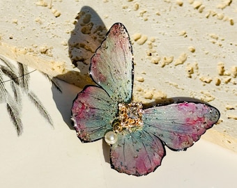 Puede incluir: Un delicado broche de mariposa con alas iridiscentes, un acento de pedrería dorada y un detalle de perla.