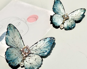 Puede incluir: Dos pinzas para el pelo con forma de mariposa azul con detalles de perlas y adornos de pedrería. Las mariposas tienen un acabado brillante e iridiscente.