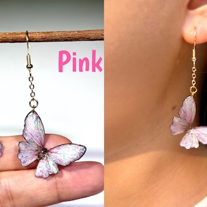K&ouml;nnte beinhalten: Rosa Schmetterlingsohrringe mit goldfarbener Kette und Haken. Die Schmetterlingsfl&uuml;gel sind zartrosa mit wei&szlig;en und schillernden Akzenten. Die Ohrringe werden an einem Ohr und auch von einer Hand gehalten gezeigt.