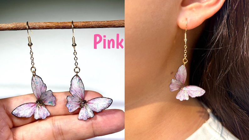K&ouml;nnte beinhalten: Rosa Schmetterlingsohrringe mit goldfarbener Kette und Haken. Die Schmetterlingsfl&uuml;gel sind zartrosa mit wei&szlig;en und schillernden Akzenten. Die Ohrringe werden an einem Ohr und auch von einer Hand gehalten gezeigt.