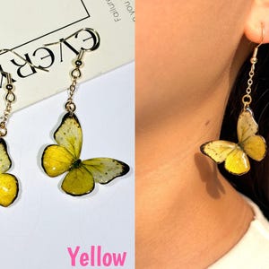 K&ouml;nnte beinhalten: Gelbe Schmetterlingsohrringe mit goldfarbenen Haken und Ketten. Die Schmetterlingsfl&uuml;gel sind gelb mit schwarzen und wei&szlig;en Details. Ein Ohrring wird von einer Person getragen. Das Wort "Yellow" ist in Pink geschrieben.