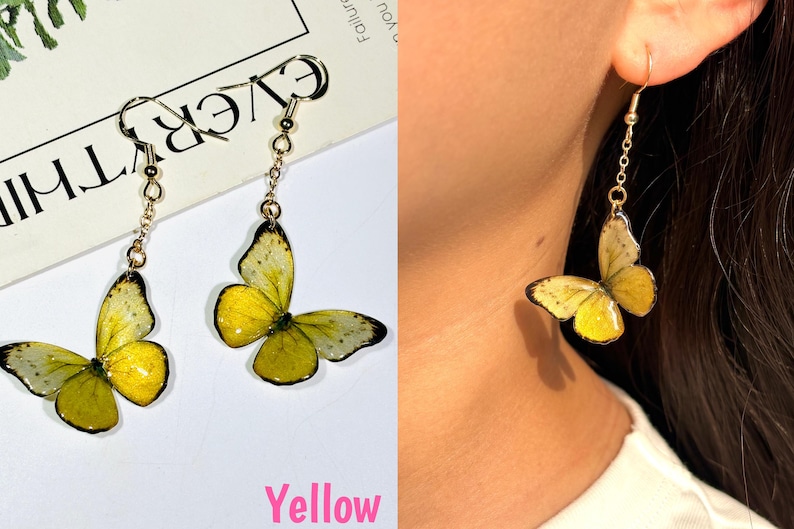 K&ouml;nnte beinhalten: Gelbe Schmetterlingsohrringe mit goldfarbenen Haken und Ketten. Die Schmetterlingsfl&uuml;gel sind gelb mit schwarzen und wei&szlig;en Details. Ein Ohrring wird von einer Person getragen. Das Wort "Yellow" ist in Pink geschrieben.
