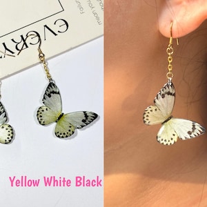 K&ouml;nnte beinhalten: Ohrringe mit Schmetterlingsanh&auml;ngern in Gelb, Wei&szlig; und Schwarz. Die Ohrringe haben eine goldfarbene Kette und einen Haken. Die Schmetterlingsfl&uuml;gel haben schwarze Punkte und Aderdetails. Der Text "Yellow White Black" ist ebenfalls im Bild enthalten.