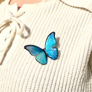 Puede incluir: Un broche de mariposa azul con detalles de purpurina fijado a un suéter de punto blanco.