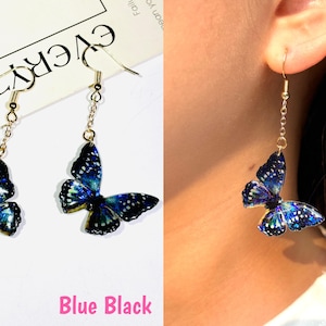 K&ouml;nnte beinhalten: H&auml;ngende Schmetterlingsohrringe mit blau-schwarzen Fl&uuml;geln. Die Schmetterlingsfl&uuml;gel haben ein detailliertes Muster und sind an einer goldfarbenen Kette und einem Haken aufgeh&auml;ngt. Der Text "Blue Black" befindet sich am unteren Rand des Bildes.