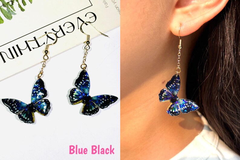 K&ouml;nnte beinhalten: H&auml;ngende Schmetterlingsohrringe mit blau-schwarzen Fl&uuml;geln. Die Schmetterlingsfl&uuml;gel haben ein detailliertes Muster und sind an einer goldfarbenen Kette und einem Haken aufgeh&auml;ngt. Der Text "Blue Black" befindet sich am unteren Rand des Bildes.