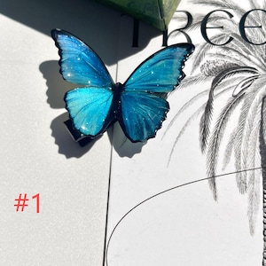 Puede incluir: Una pinza para el pelo con forma de mariposa azul con detalles de purpurina iridiscente. La mariposa tiene patrones de alas detallados y est&aacute; sujeta a una pinza negra. El n&uacute;mero "#1" est&aacute; impreso en rojo en el fondo.
