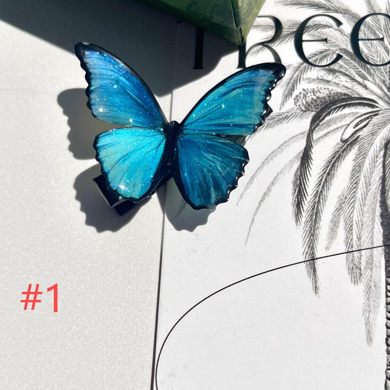 Puede incluir: Una pinza para el pelo con forma de mariposa azul con detalles de purpurina iridiscente. La mariposa tiene patrones de alas detallados y est&aacute; sujeta a una pinza negra. El n&uacute;mero "#1" est&aacute; impreso en rojo en el fondo.