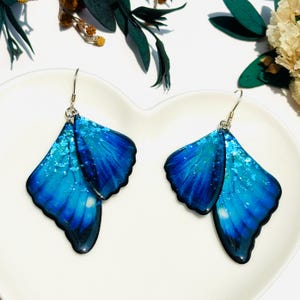 Puede incluir: Un par de pendientes de mariposa azul con ganchos plateados. Las alas de la mariposa est&aacute;n superpuestas y tienen un acabado brillante.
