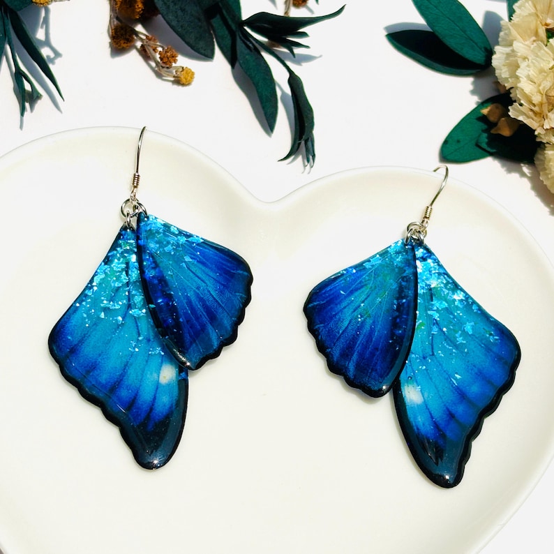 Puede incluir: Un par de pendientes de mariposa azul con ganchos plateados. Las alas de la mariposa est&aacute;n superpuestas y tienen un acabado brillante.