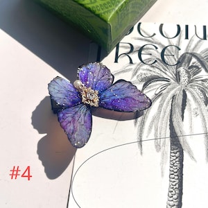 Puede incluir: Una pinza para el pelo con forma de mariposa en tonos morados y azules, con detalles dorados y una joya central. La pinza tiene una perla y detalles de purpurina. El n&uacute;mero "#4" es visible en la esquina inferior izquierda.