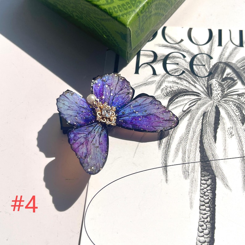 Puede incluir: Una pinza para el pelo con forma de mariposa en tonos morados y azules, con detalles dorados y una joya central. La pinza tiene una perla y detalles de purpurina. El n&uacute;mero "#4" es visible en la esquina inferior izquierda.