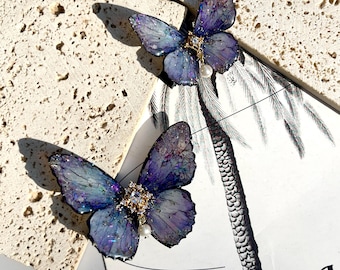 May include: A pair of iridescent blue butterfly earrings with a pearl and rhinestone accent. The butterflies are set against a white background.
