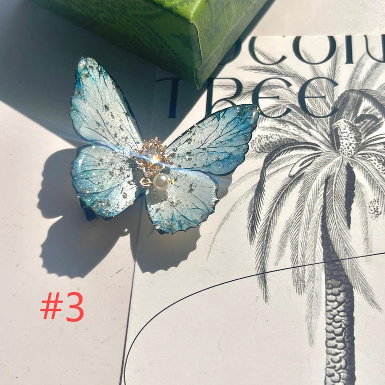 Puede incluir: Una pinza para el pelo con forma de mariposa azul y blanca con detalles de purpurina y una perla en el centro. La pinza tiene un adorno dorado y est&aacute; sobre un fondo blanco con una ilustraci&oacute;n de palmera. El n&uacute;mero "#3" es visible en rojo.