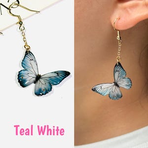 K&ouml;nnte beinhalten: T&uuml;rkis-wei&szlig;e Schmetterlingsohrringe mit goldfarbenen Haken und Ketten. Die Schmetterlingsfl&uuml;gel haben ein detailliertes Design mit t&uuml;rkisfarbener und wei&szlig;er Farbe. Ein Ohrring wird getragen.