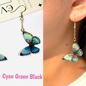 K&ouml;nnte beinhalten: Ohrringe mit Schmetterlingen in Cyan, Gr&uuml;n und Schwarz. Die Ohrringe haben eine goldfarbene Kette und einen Haken. Die Schmetterlingsfl&uuml;gel haben ein detailliertes Muster und sind an einer goldfarbenen Kette befestigt.